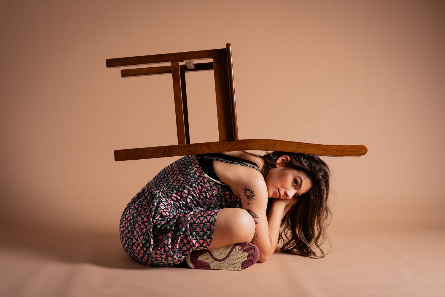 Artistic pose with wooden chair and patterned dress against warm studio backdrop.
