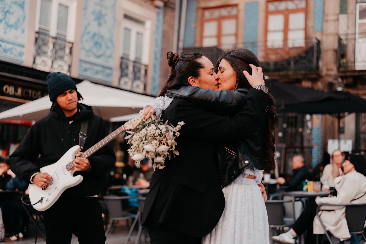 Two women share a kiss in a vibrant plaza while a guitarist performs nearby, with blue tiled buildings in the background.