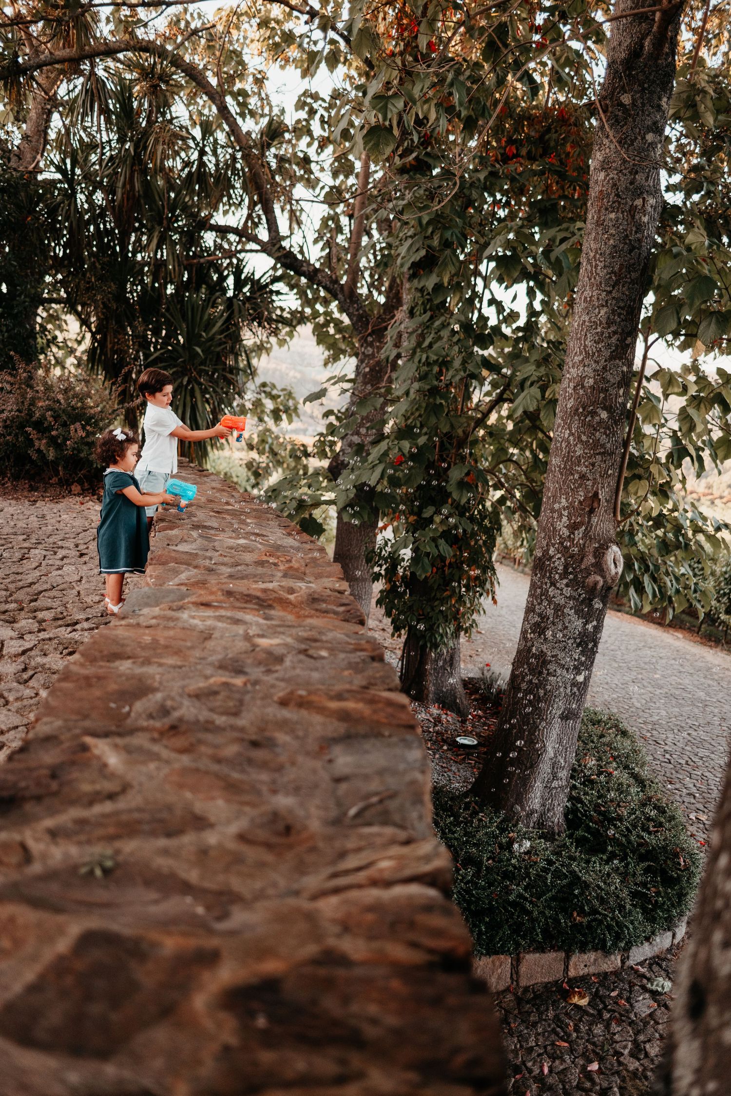 A woman and child play with colorful water guns on a stone wall path surrounded by lush green trees.