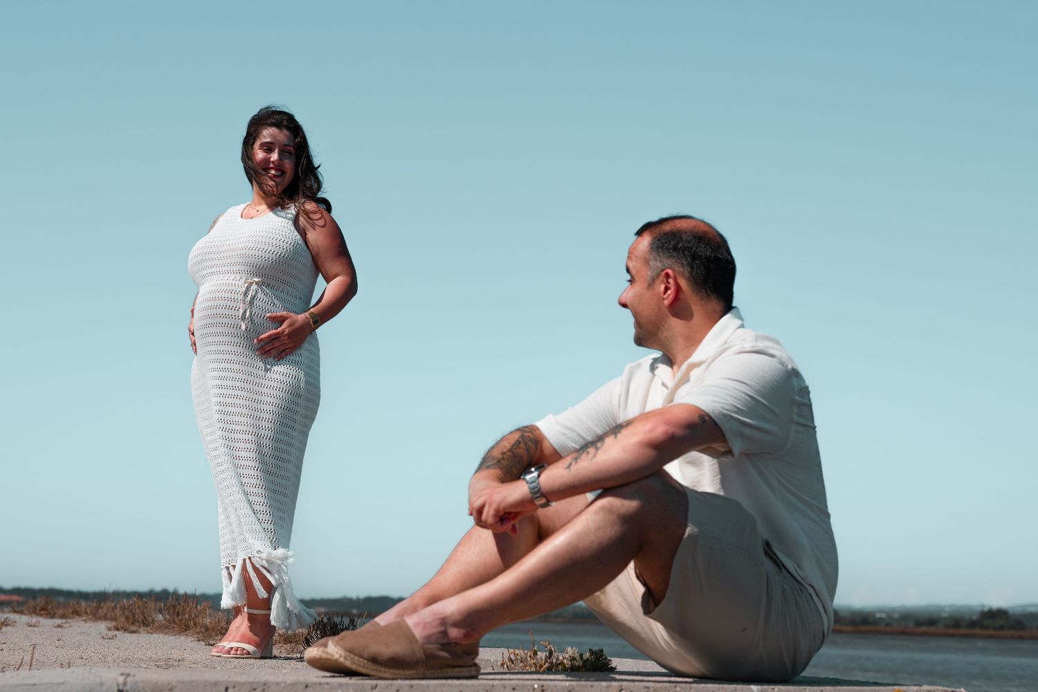 Couple in matching white outfits posing on a concrete ledge against blue sky.