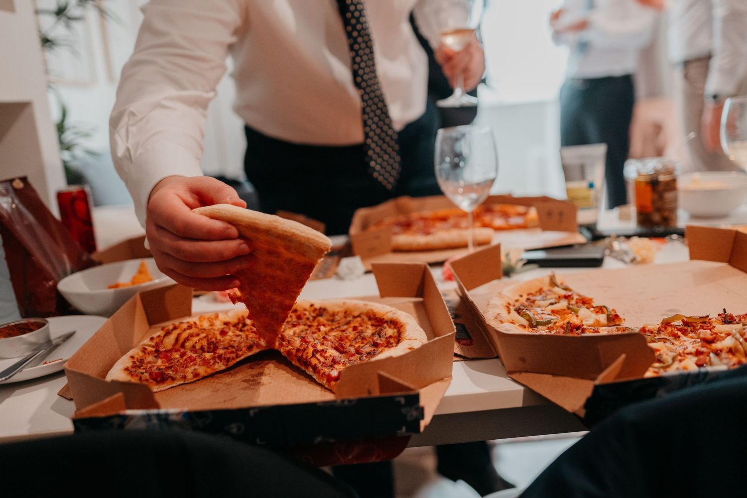 People serving pizza slices from open cardboard boxes at a catered business event with glasses on the table.