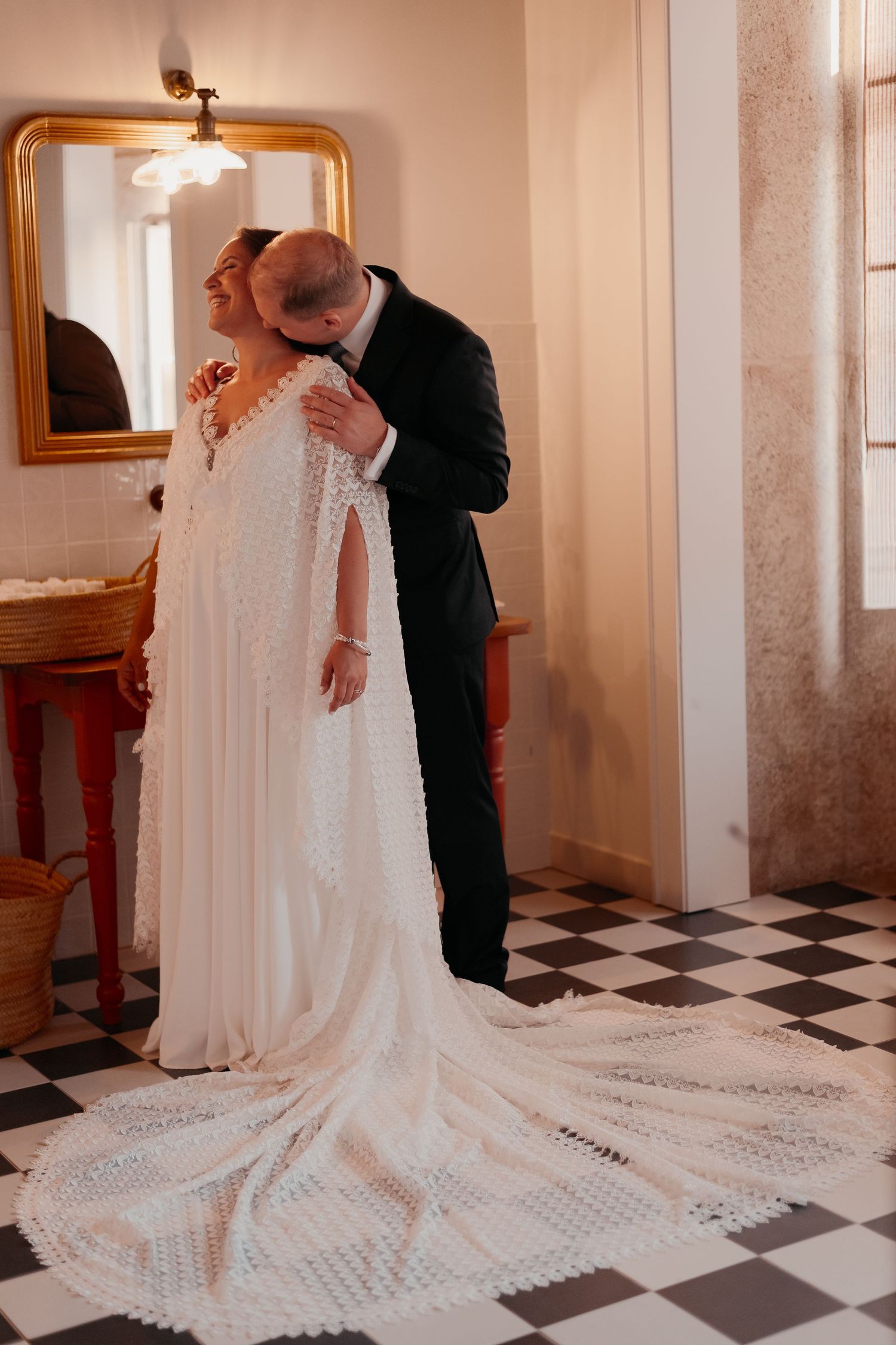 Groom in black tuxedo kissing bride in elegant white lace cape wedding dress on checkered floor.