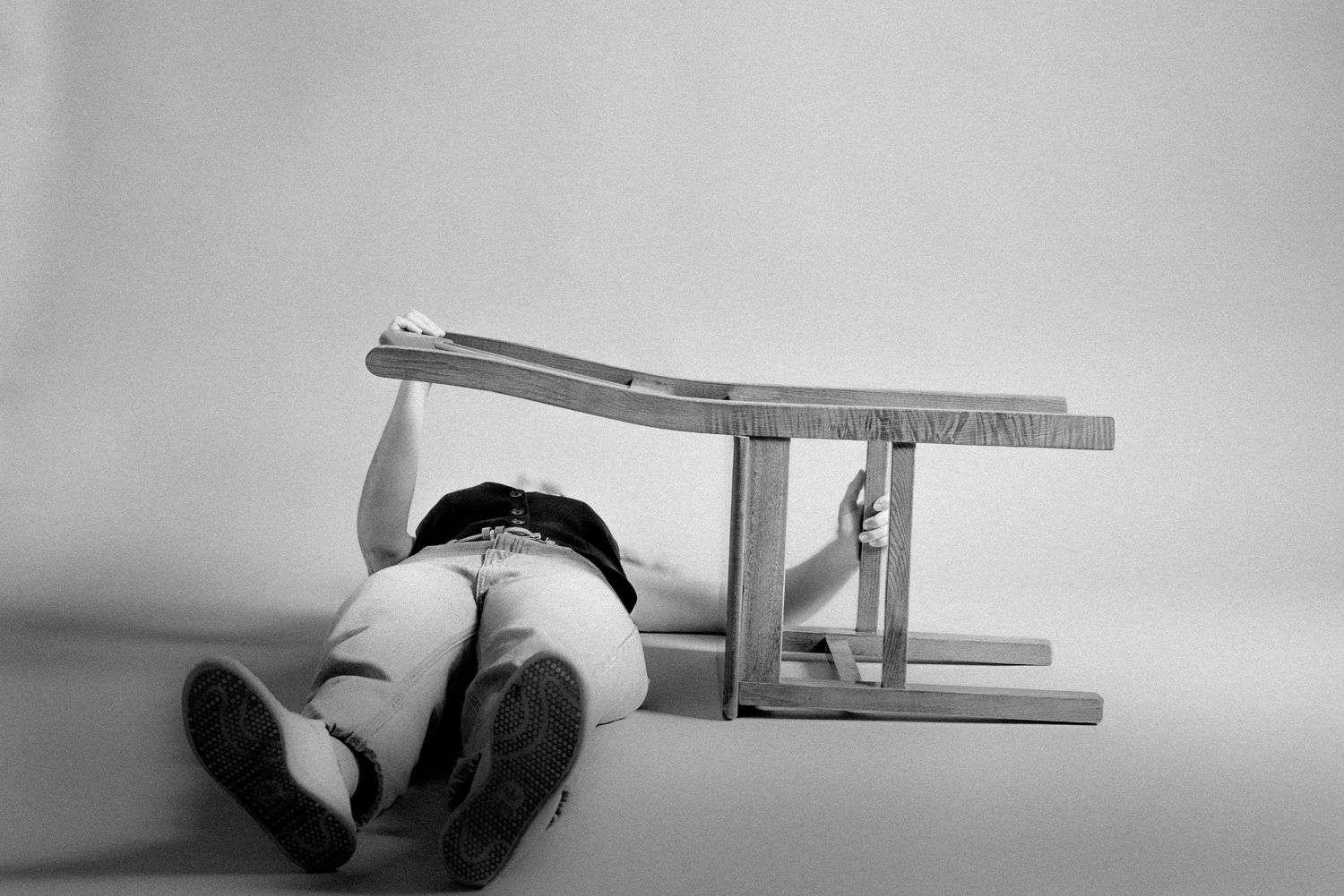 Black and white image of someone lying under an overturned wooden chair.