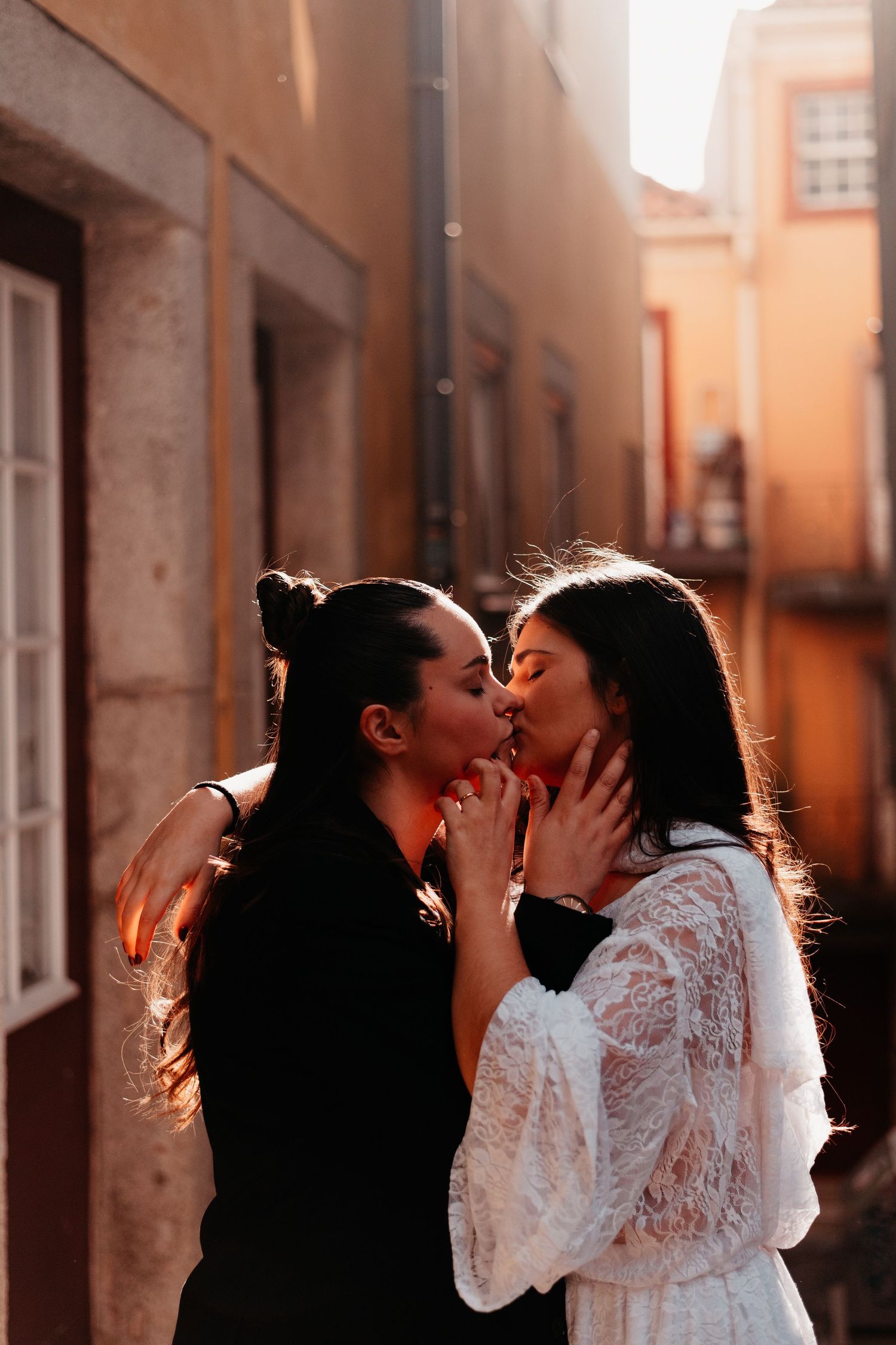Two women sharing a romantic kiss in a sunlit alley between old buildings.