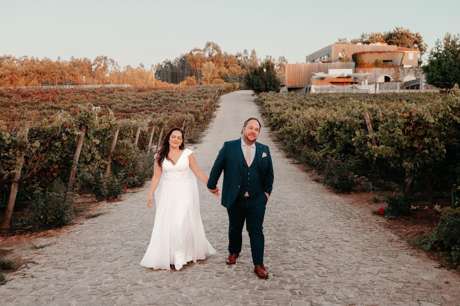 Bride and groom holding hands on a vineyard path at sunset, surrounded by lush green vines and autumn foliage.