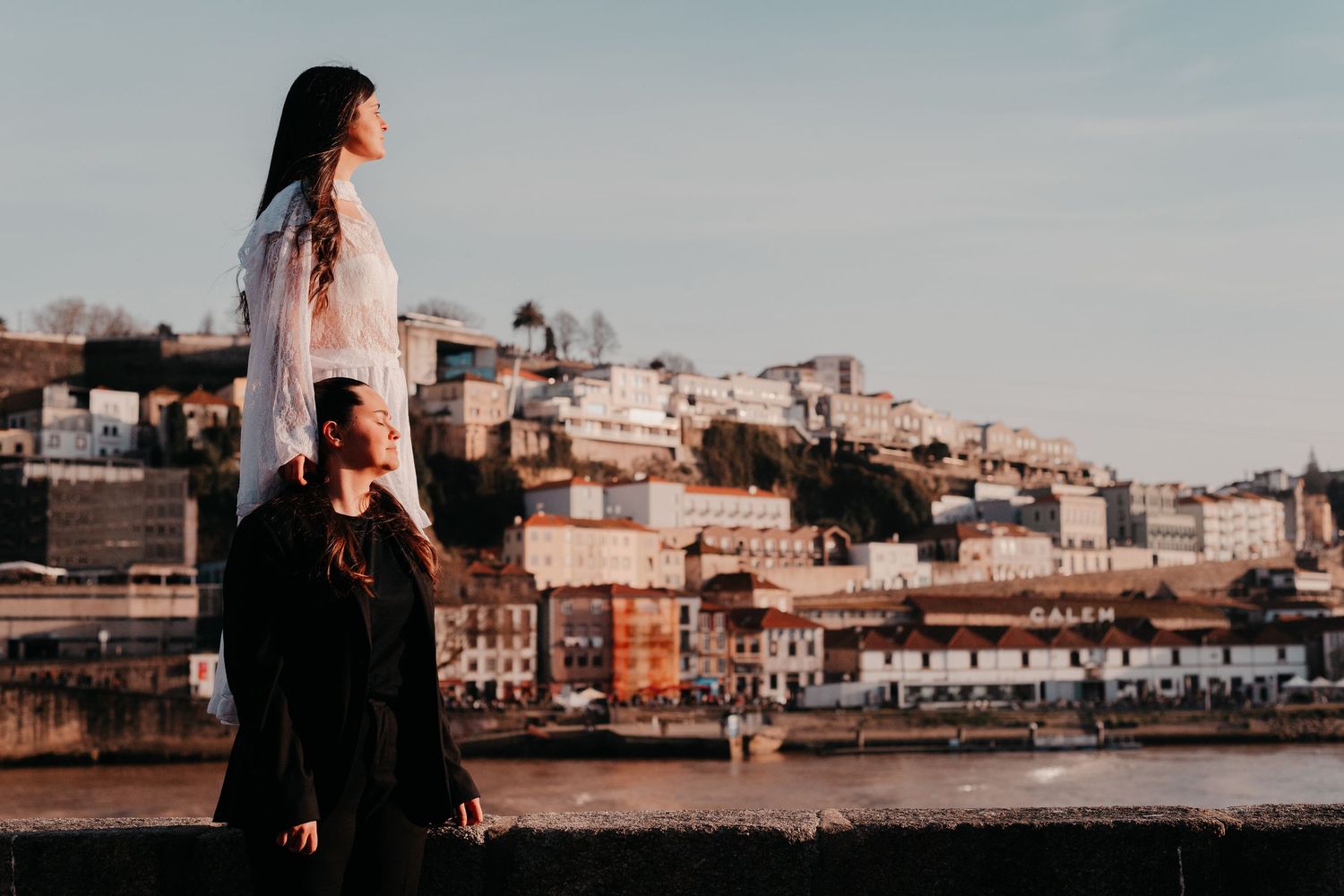 Couple standing on a riverside wall with Porto cityscape and golden sunset light in the background.