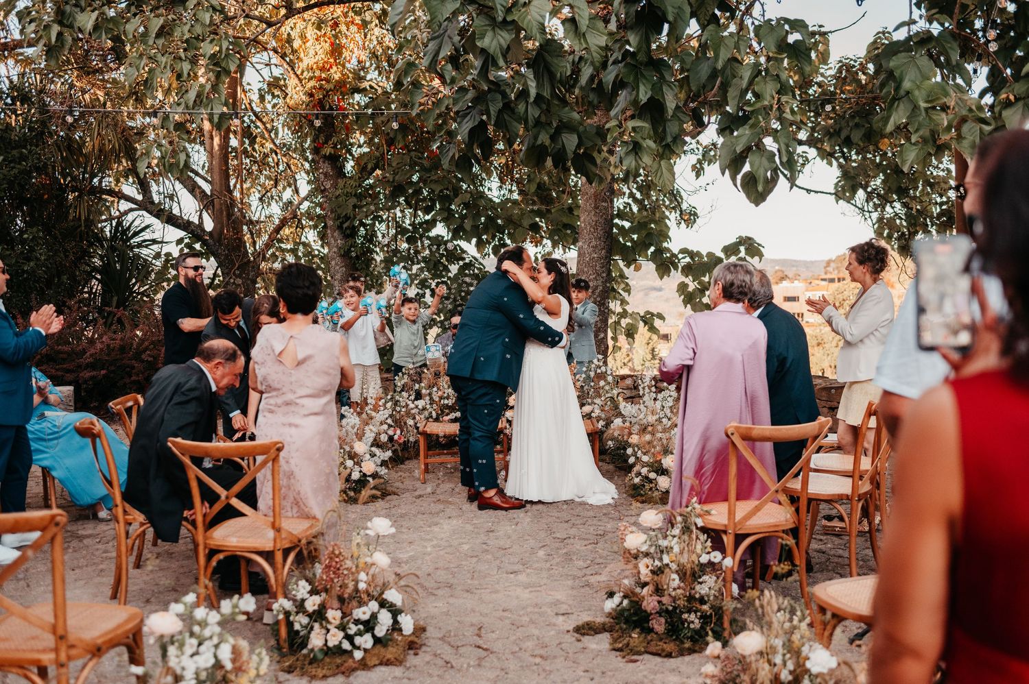 Couple shares first kiss at outdoor wedding ceremony under tree with floral arrangements and guests applauding.