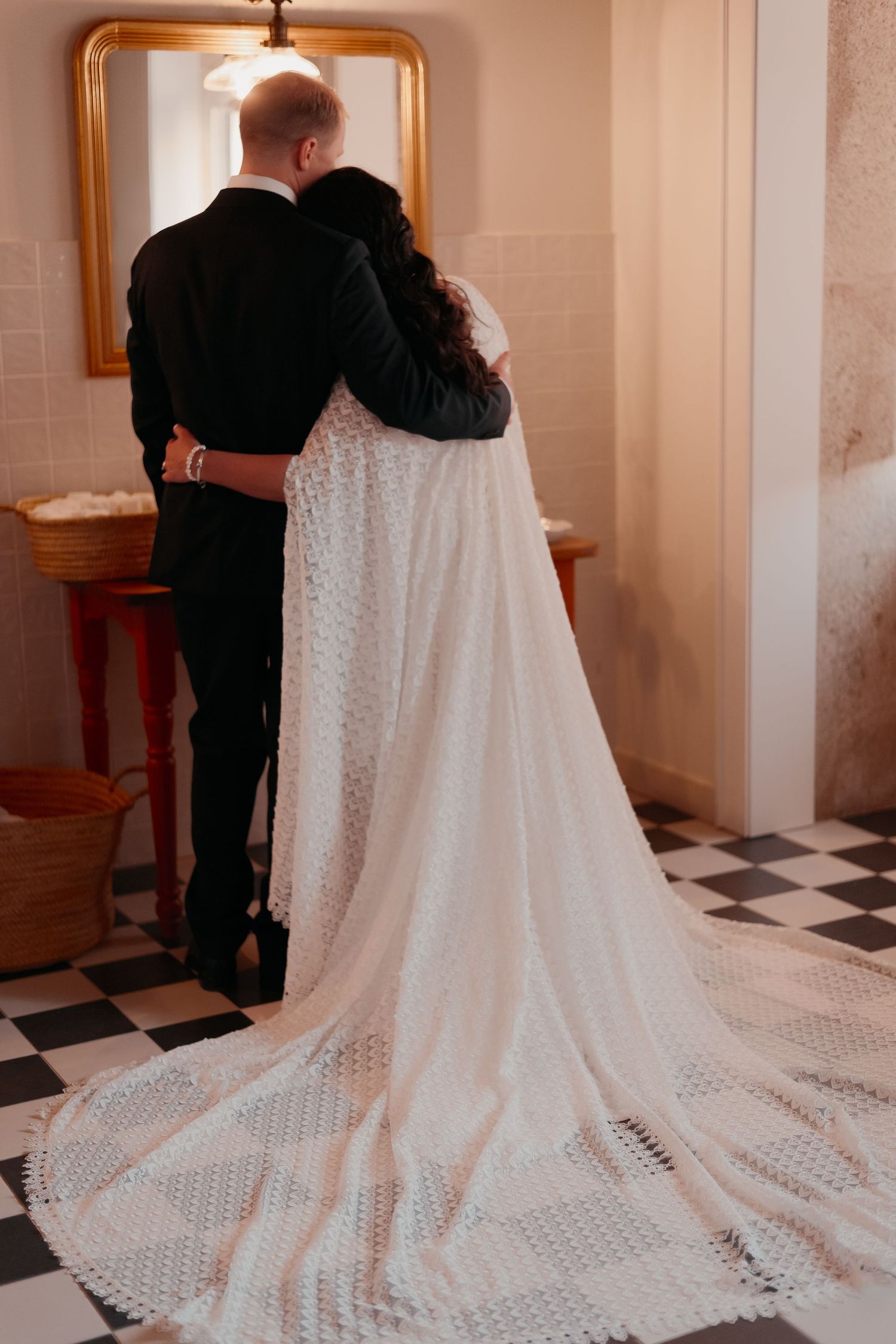 Bride and groom embracing from behind in elegant wedding attire, bride wearing lace train gown on checkered floor.