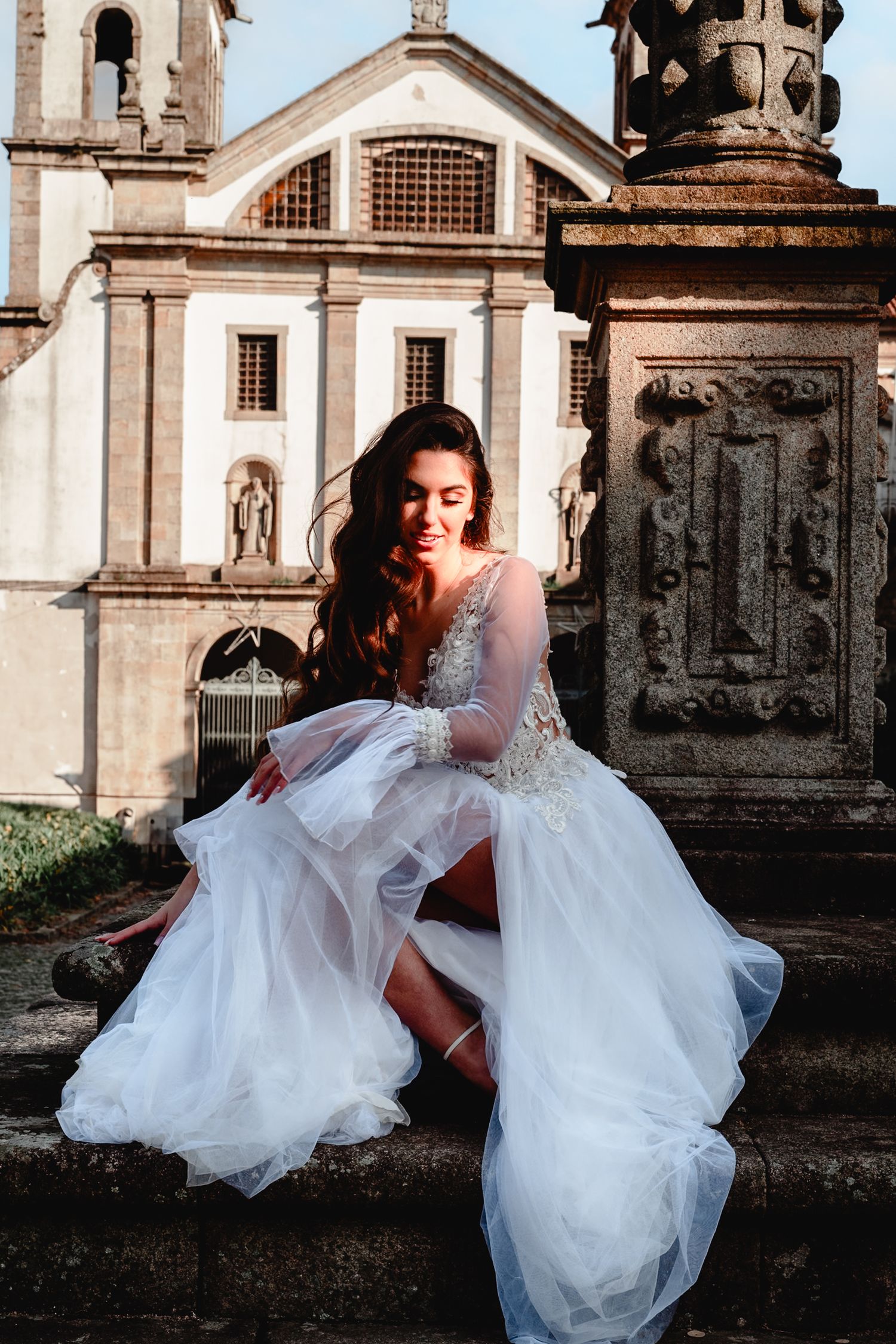 Elegant bride in detailed white gown poses on stone steps of historic church architecture.