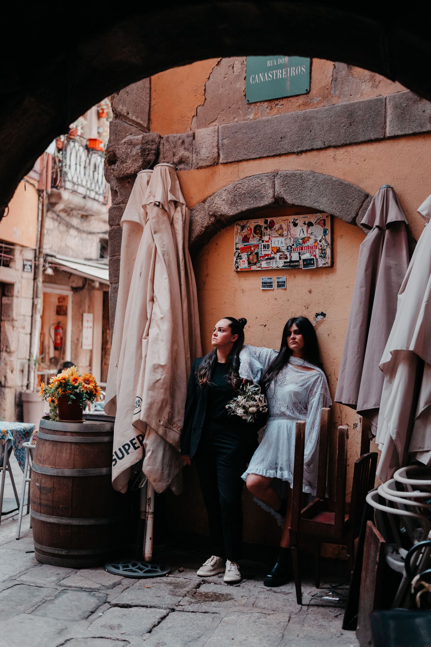 Two women posing under a stone archway at a charming outdoor European cafe with wine barrels and umbrellas.