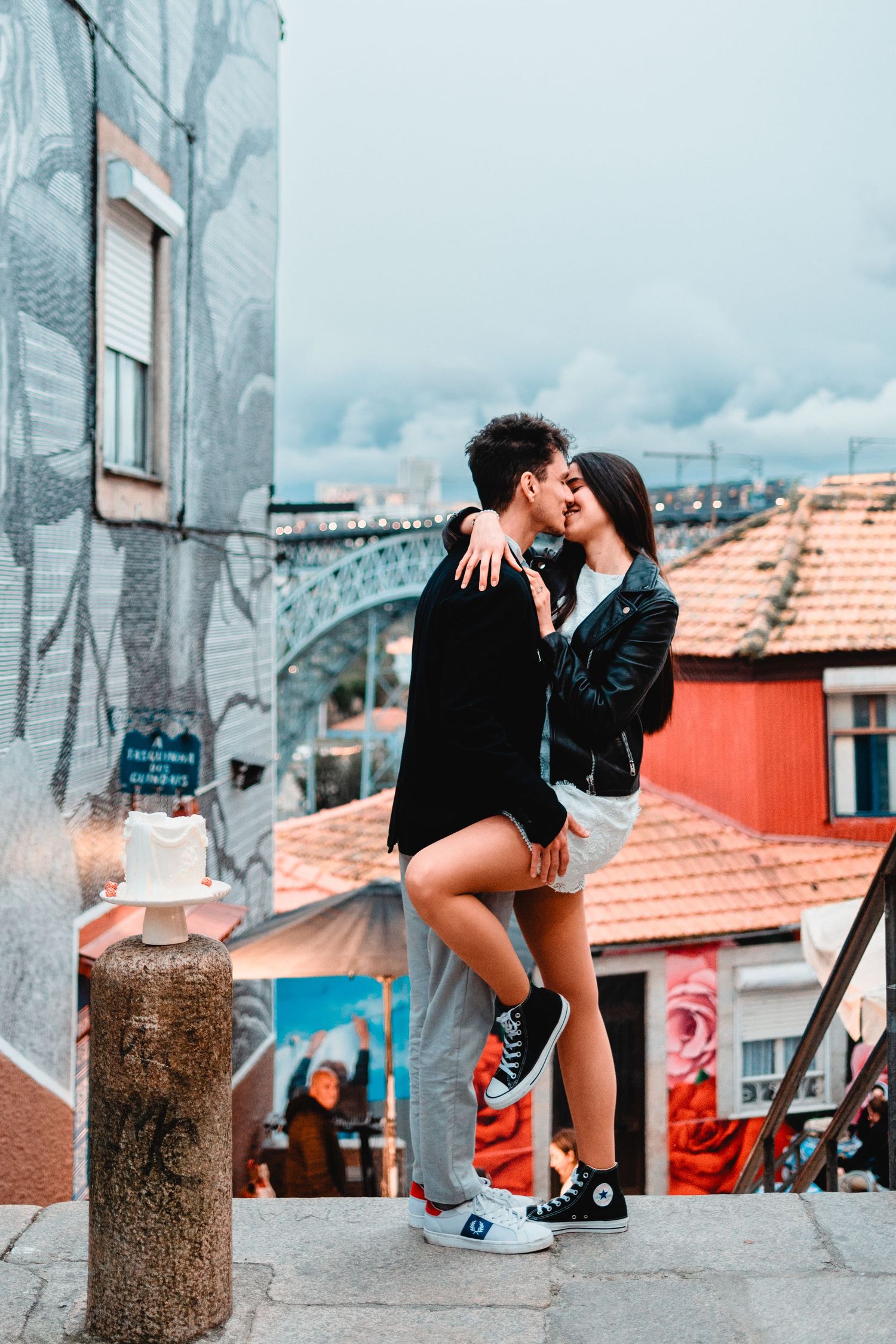 Couple sharing a romantic kiss on urban steps with colorful street art and red rooftops in the background.