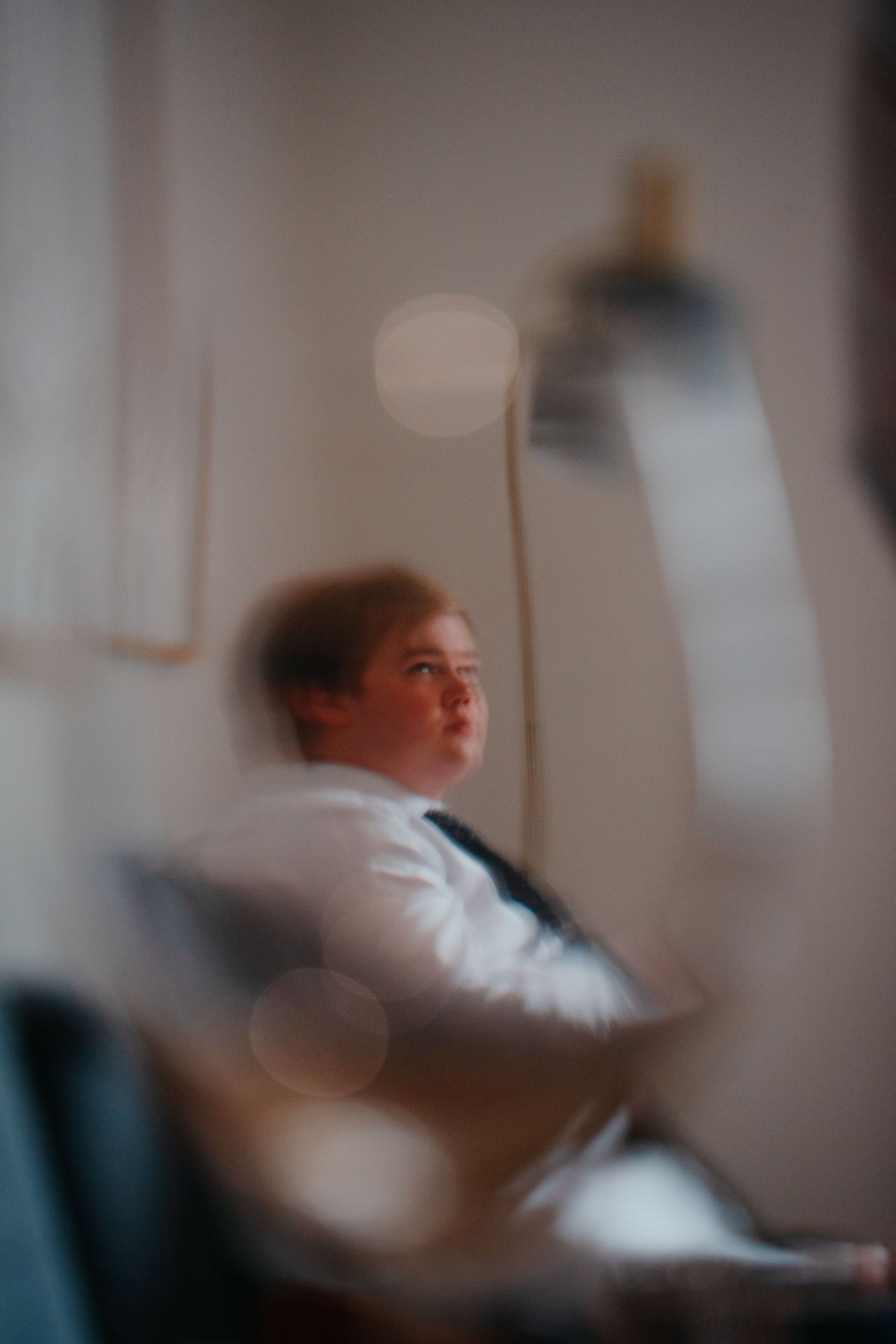 A young boy in a white shirt sits thoughtfully in a chair, captured in a softly blurred, moody indoor portrait.