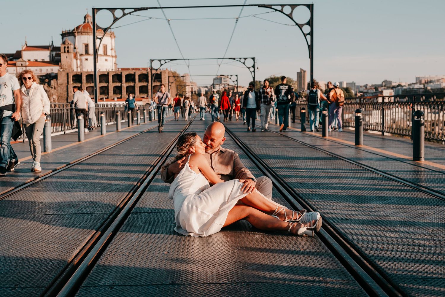 Couple sitting romantically on tram tracks of a busy bridge in Porto, Portugal, with crowds walking nearby.