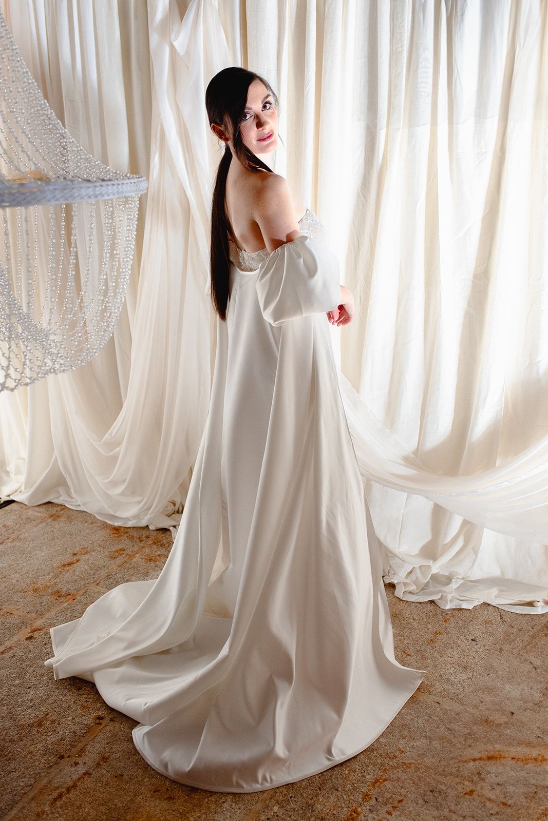 Elegant floor-length white wedding dress with off-shoulder design and flowing train against white curtain backdrop.