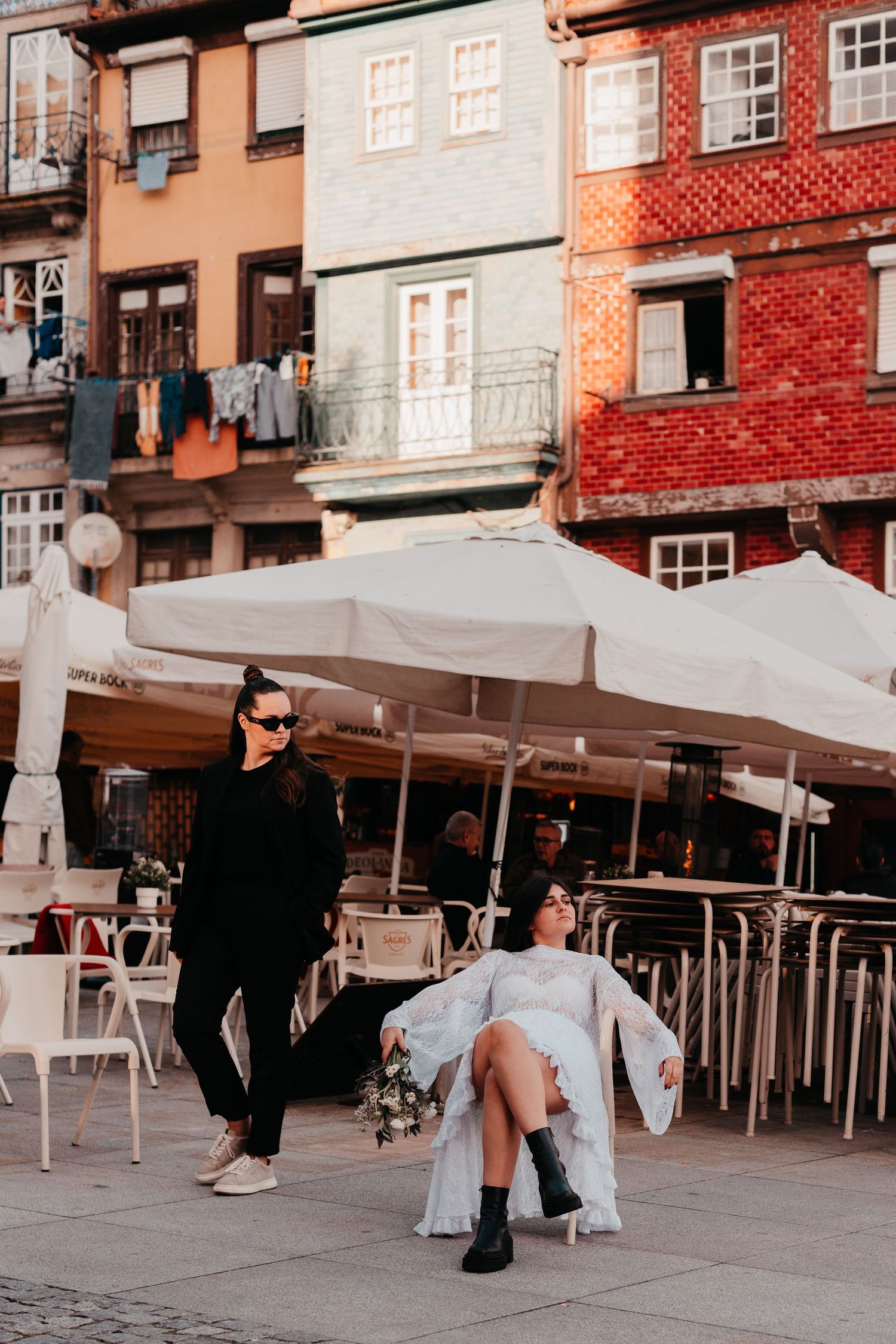 Two women in sunglasses pose near outdoor cafe tables with colorful European buildings in the background.
