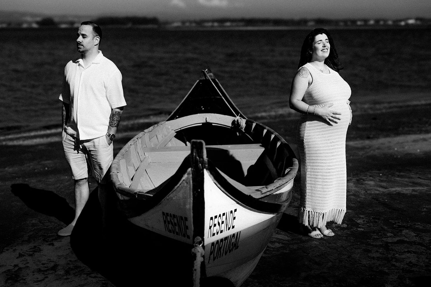 Black and white photo of a pregnant couple standing beside a wooden boat on a beach at sunset.