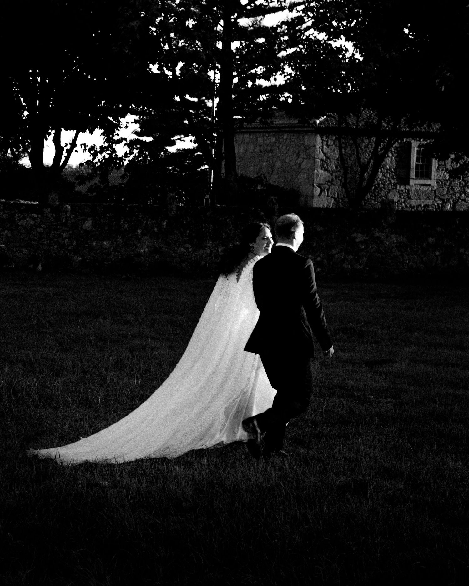 Black and white wedding photo of bride and groom walking across lawn at dusk, long veil trailing behind.