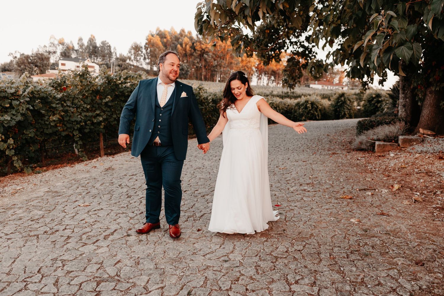 Bride and groom holding hands walking on cobblestone path, bride in white gown, groom in navy suit, outdoor wedding photo.