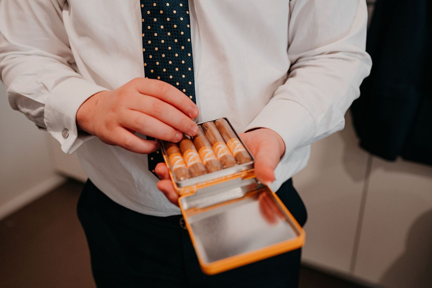 Man in white shirt and polka dot tie opening a yellow cigarette case filled with cigars.