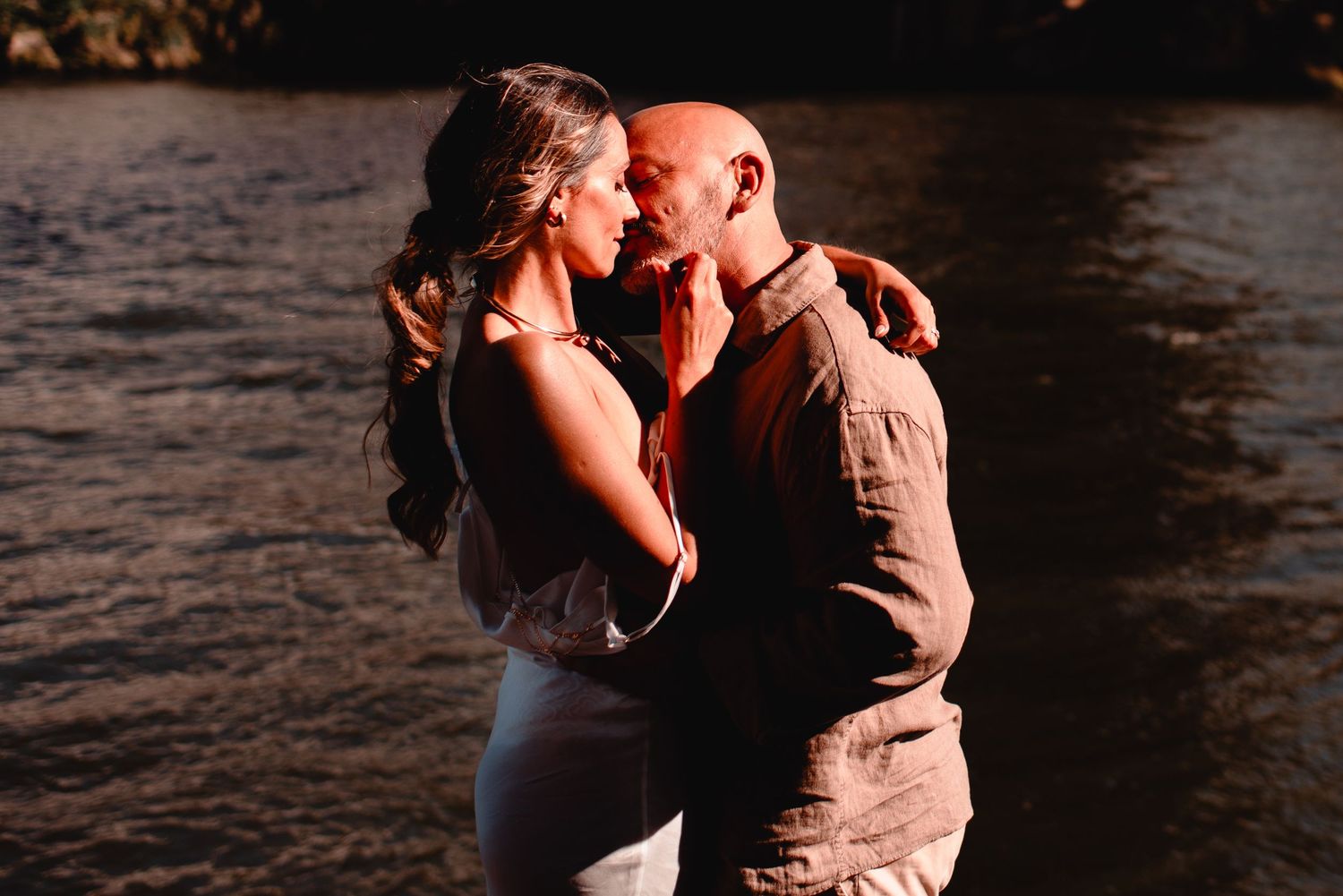 Silhouetted couple embracing near water during golden hour with warm sun flares.