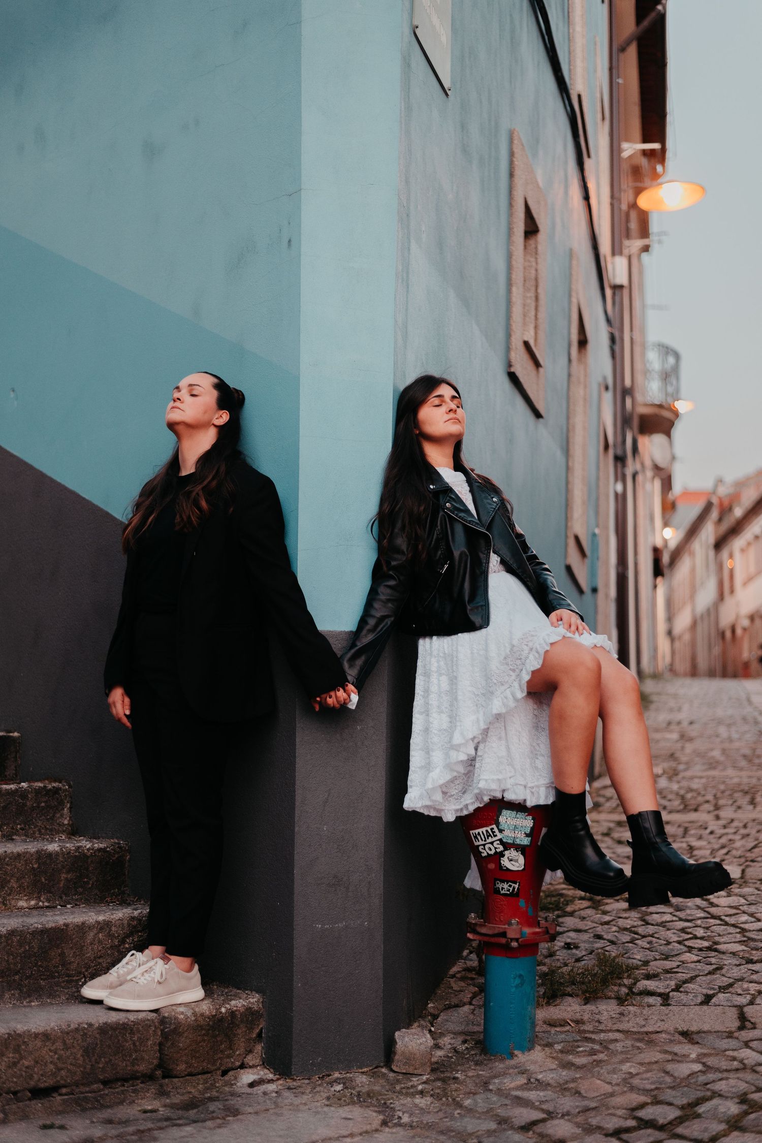 Two young women in black jackets pose against a teal building wall on a cobblestone street.