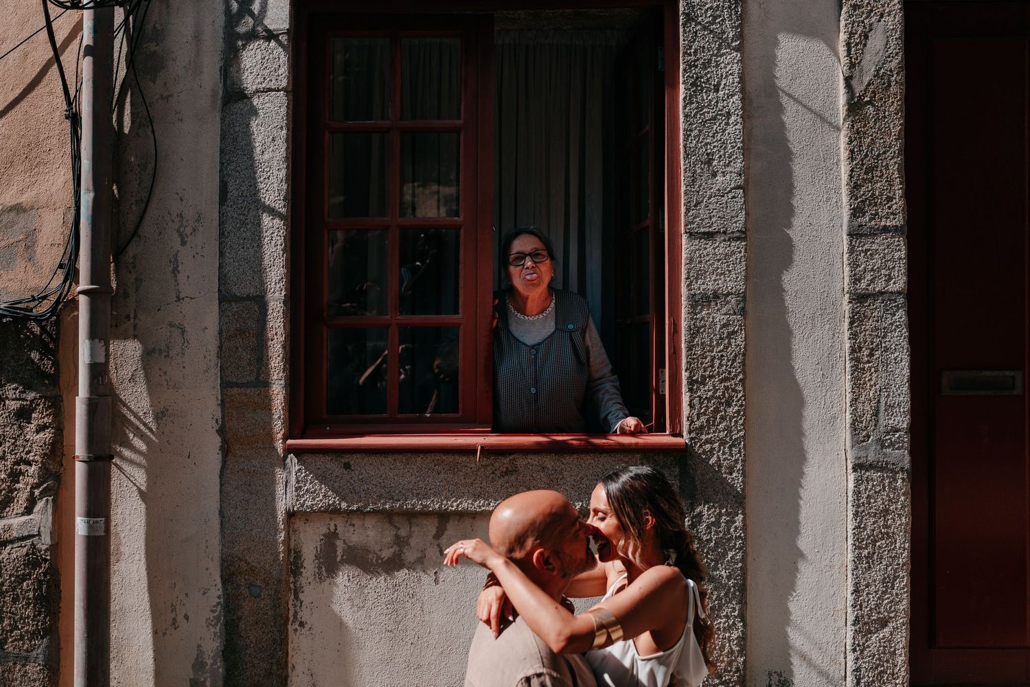 Couple embracing outside a stone building while a woman watches from a red-framed window above in Porto.