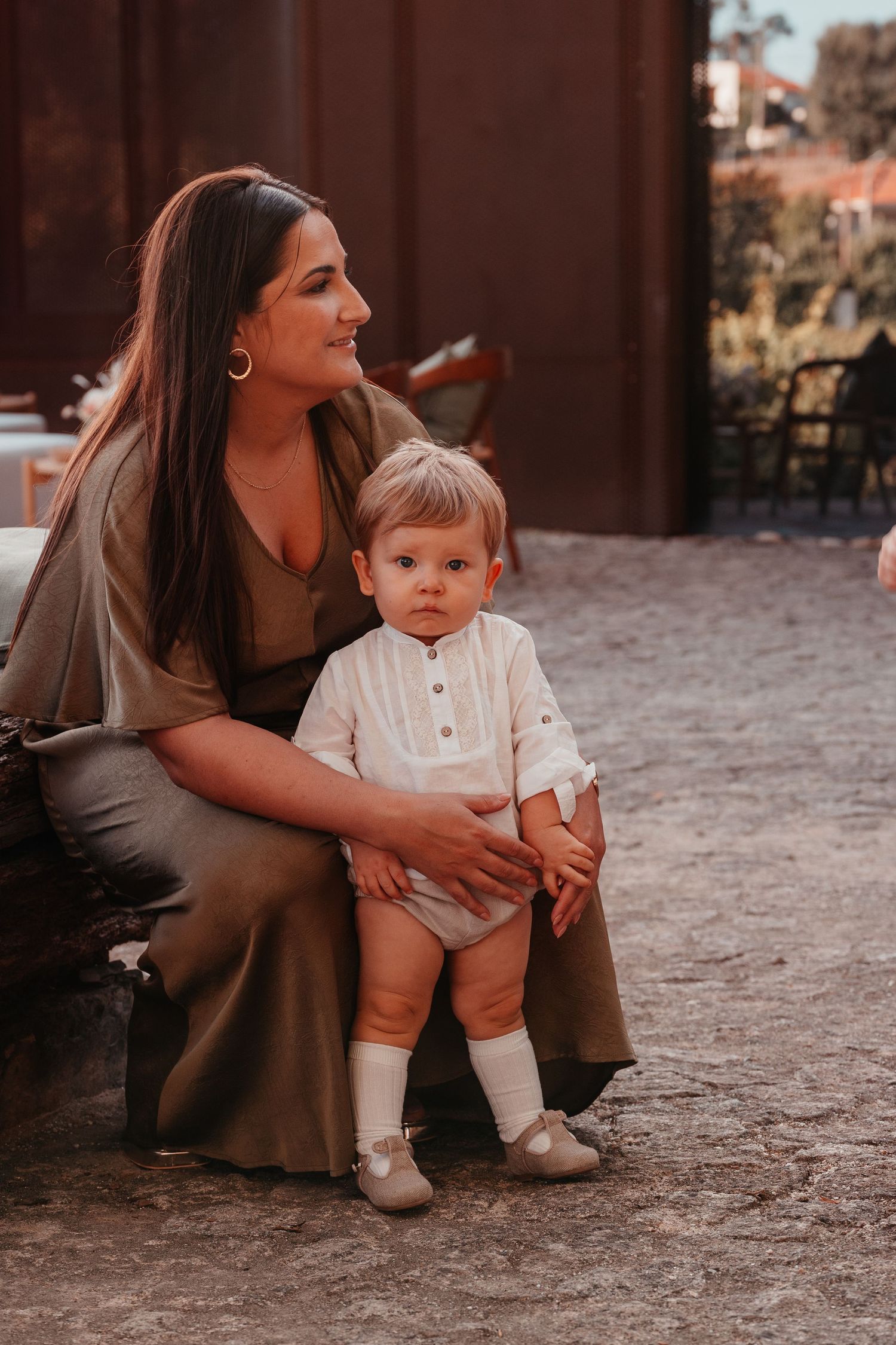 A young mother in an olive dress sits with her toddler dressed in white on a rustic cobblestone outdoor setting.