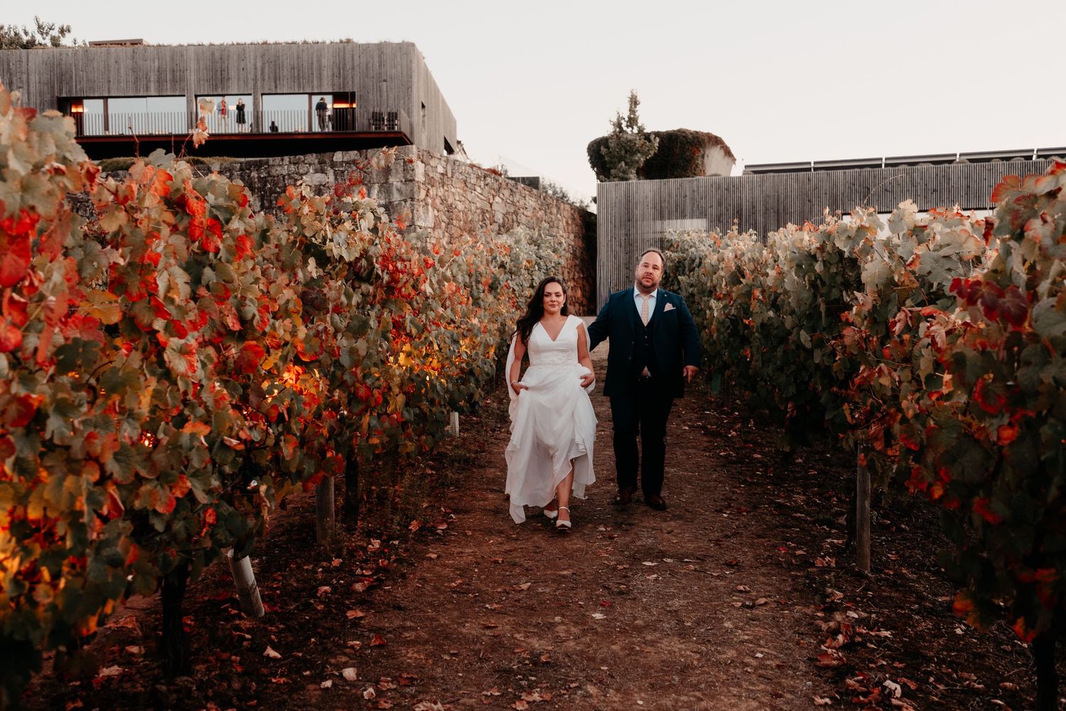 Bride and groom walk hand-in-hand through vibrant autumn vineyard rows with colorful red and orange foliage.