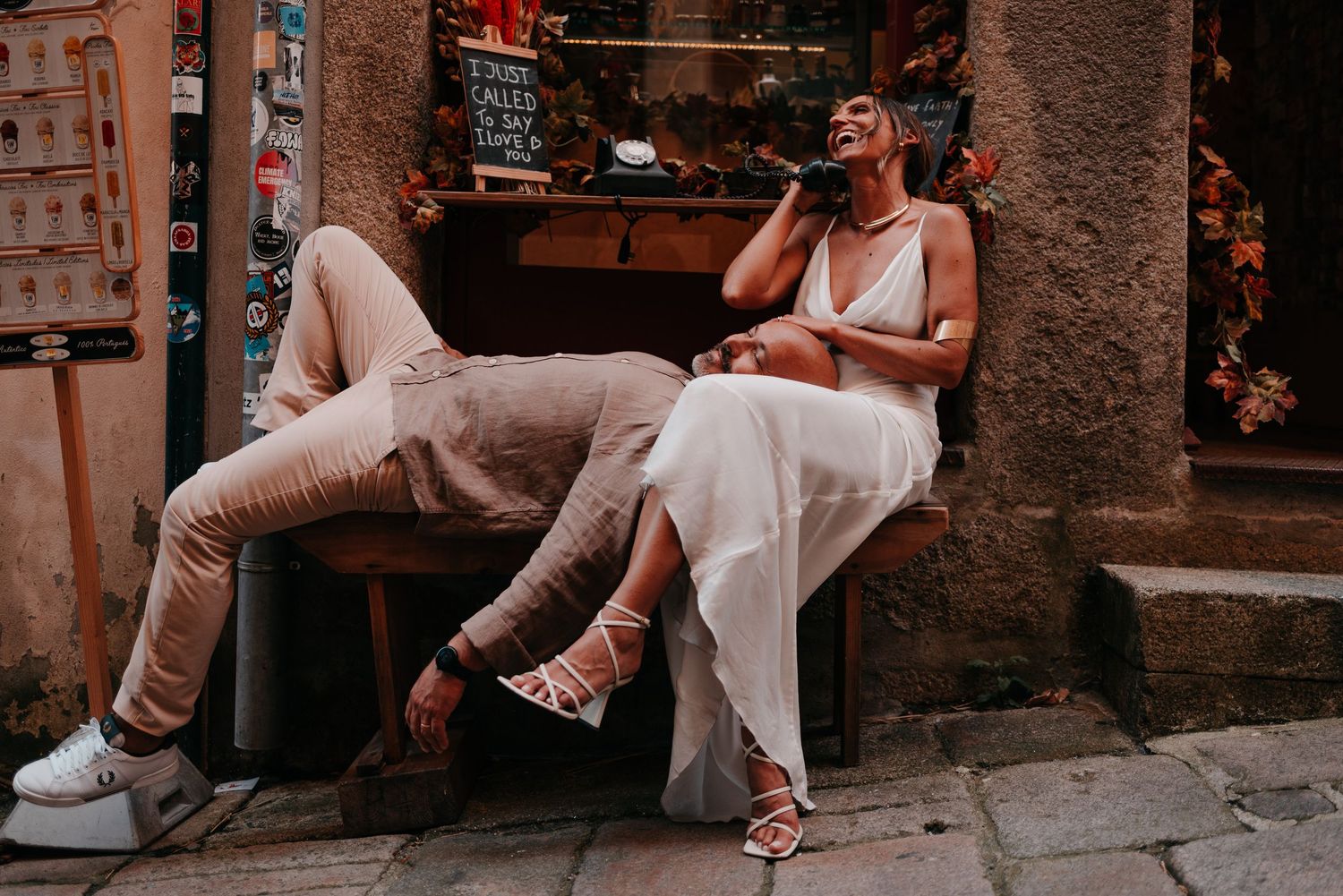 Bride and groom relaxing on a bench outside a charming Italian shop, laughing together on their wedding day.