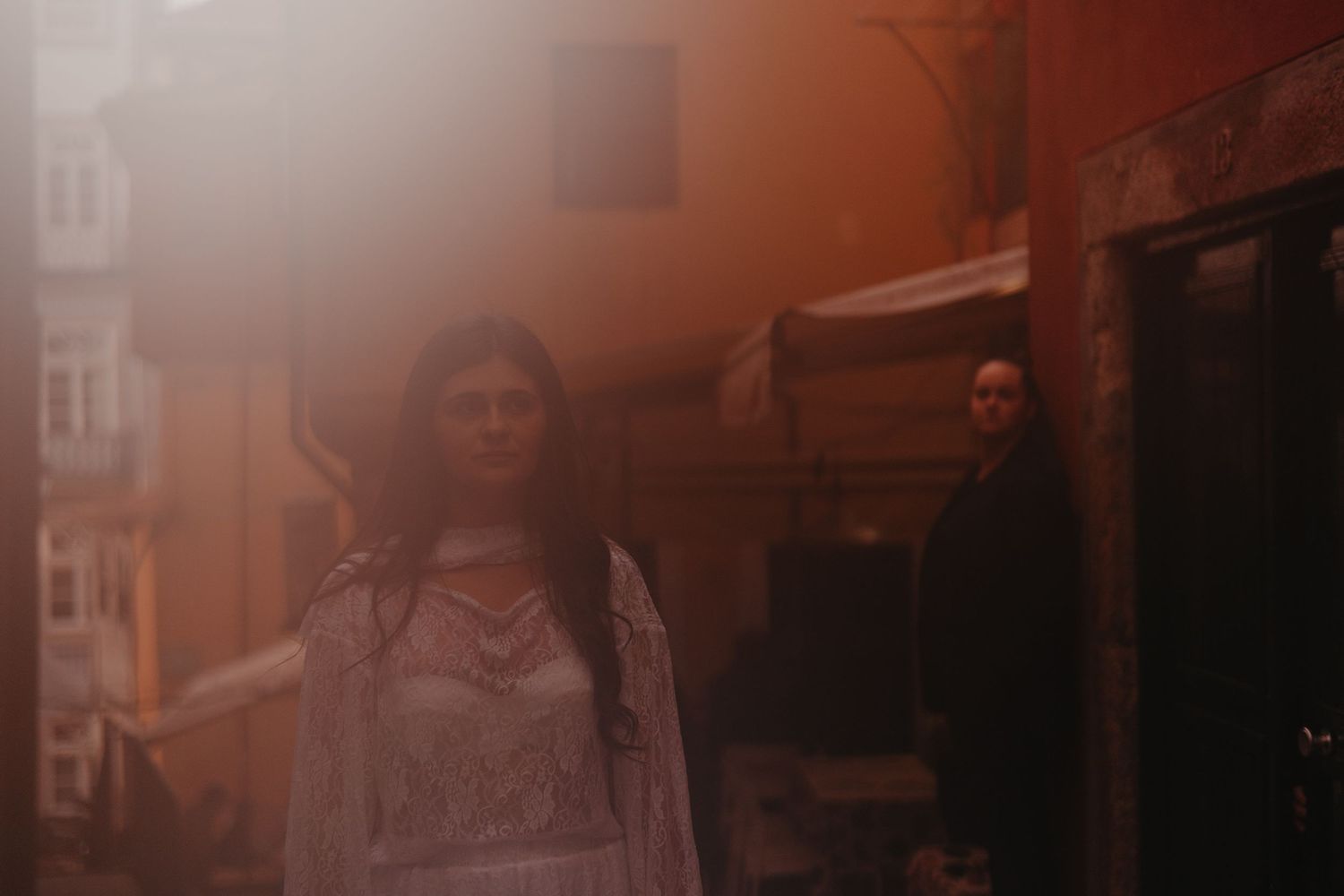 Woman in white embroidered dress stands in warm golden light on a city street with a figure in background.