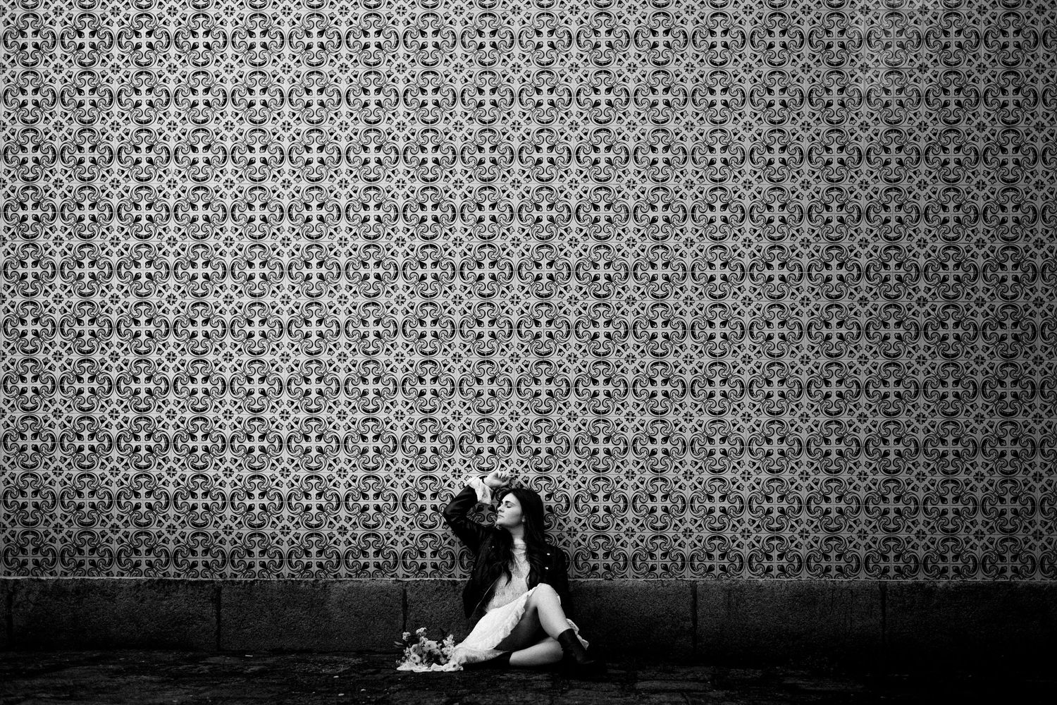 Black and white photo of a woman sitting against an ornate patterned tile wall, looking upward dramatically.