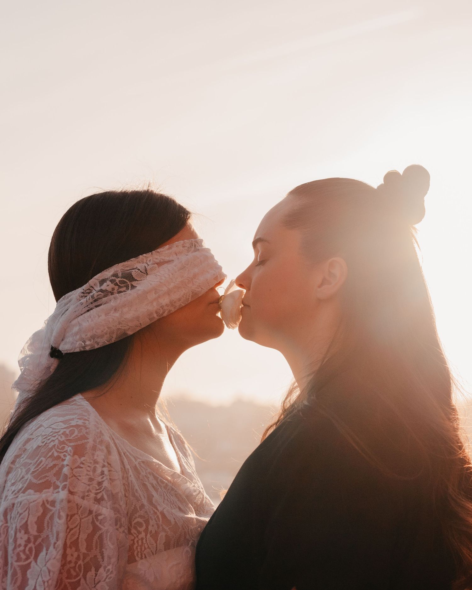 Two women sharing a romantic kiss at sunset, one wearing a lace blindfold, backlit by golden hour light.