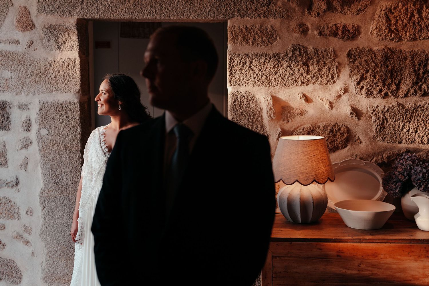 Bride in white lace gown stands in stone doorway as groom silhouette poses in foreground at rustic venue.