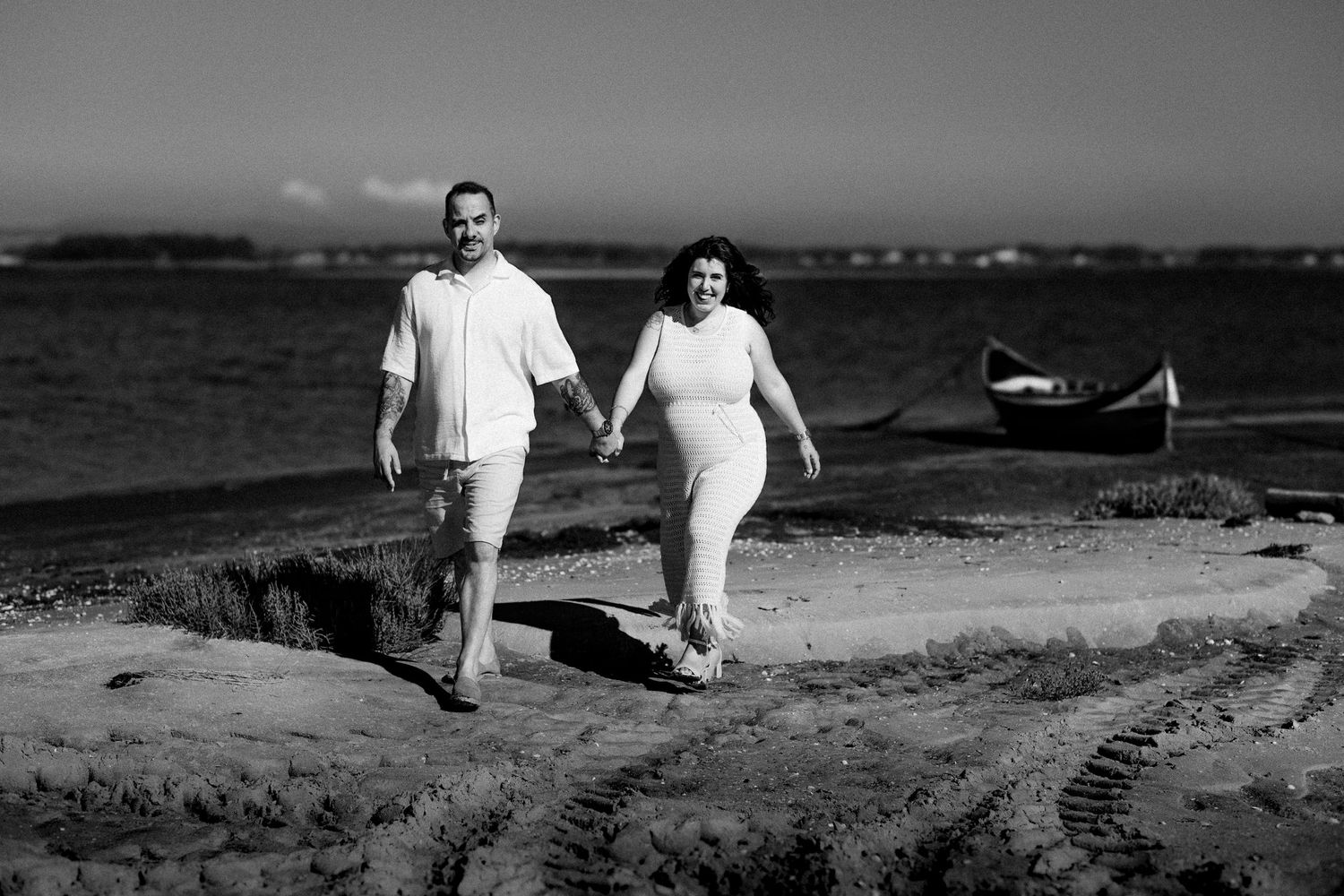 Black and white photo of couple walking hand in hand on beach with boat in background.