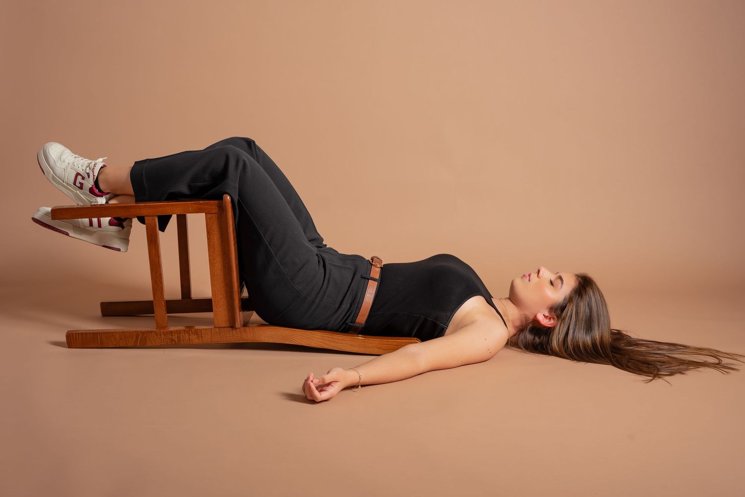 Model laying on wooden chair against peach colored backdrop wearing dark outfit and white sneakers.