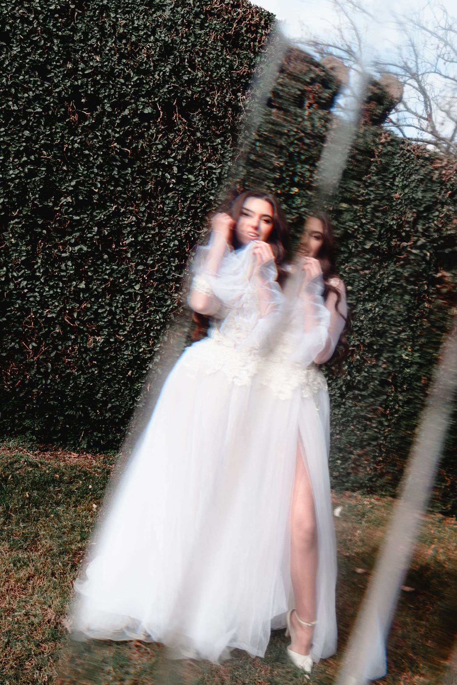 Dreamy double exposure of bride in white gown against garden hedge.