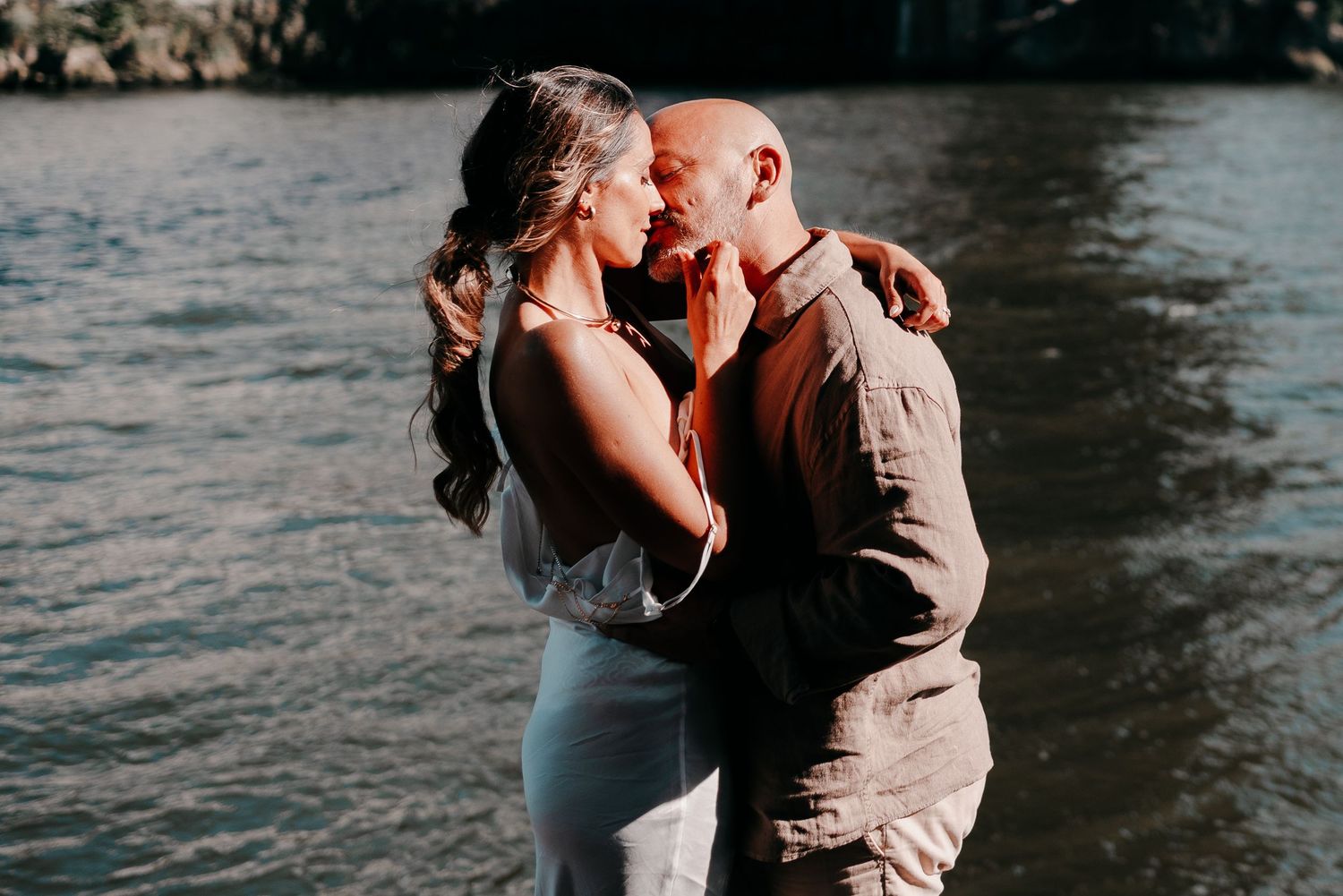 Romantic couple embracing near water, sharing an intimate moment outdoors in warm golden light.