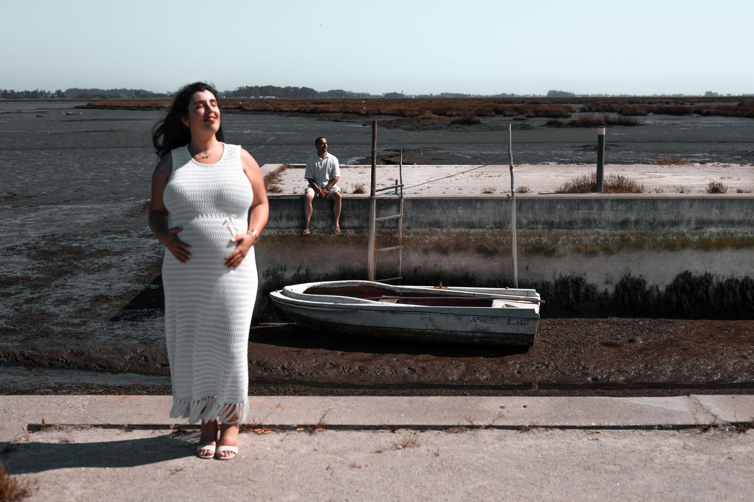 Woman in white dress standing near docked boat on waterfront.