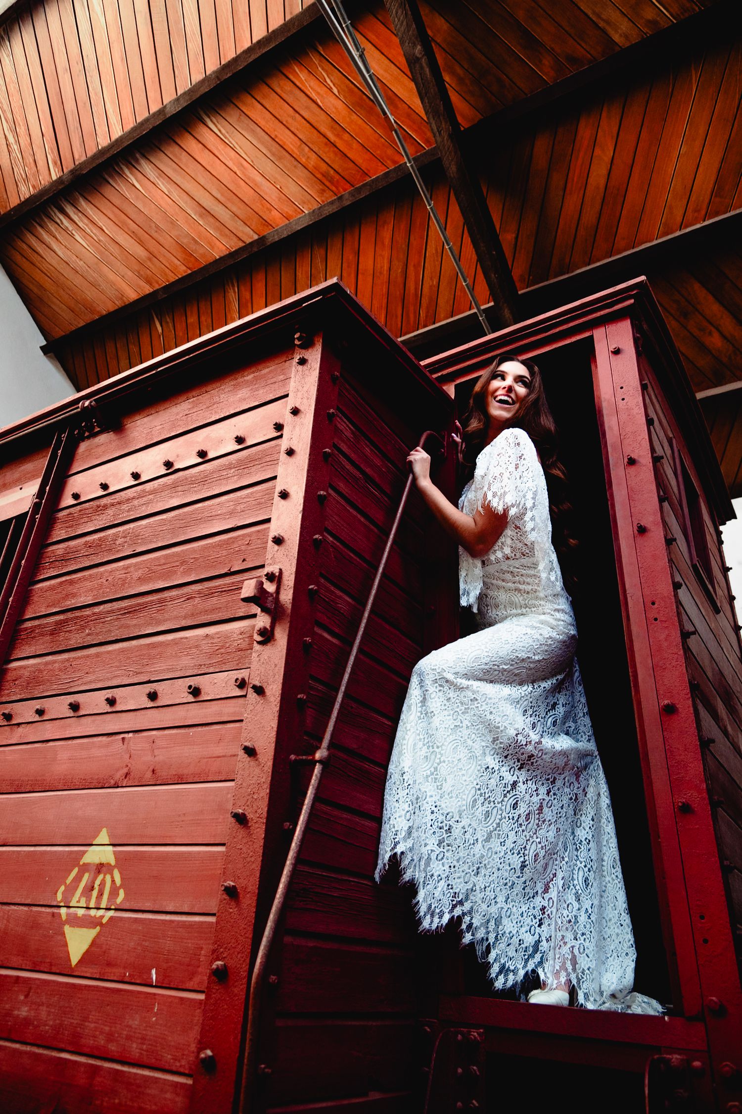 Dramatic upward angle of bride in lace gown posing in vintage red train car doorway.