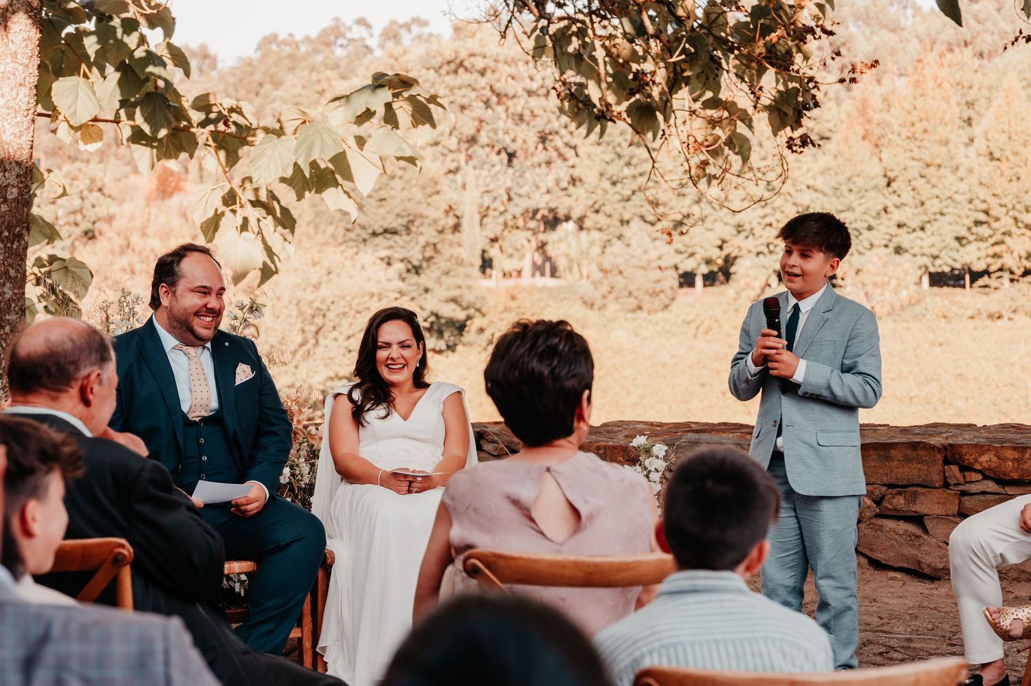Man in gray suit speaks at outdoor wedding ceremony while smiling couple sits nearby under autumn trees.