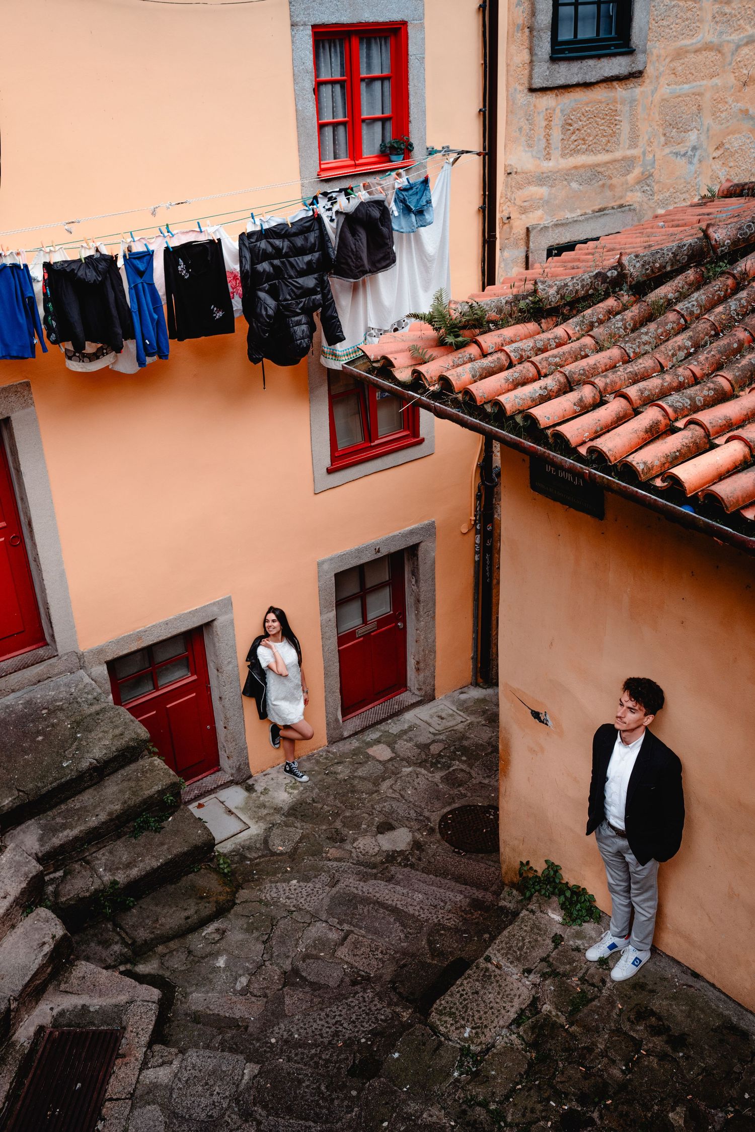 Couple posing in a charming European alley with colorful buildings, red windows, and laundry hanging above cobblestone steps.