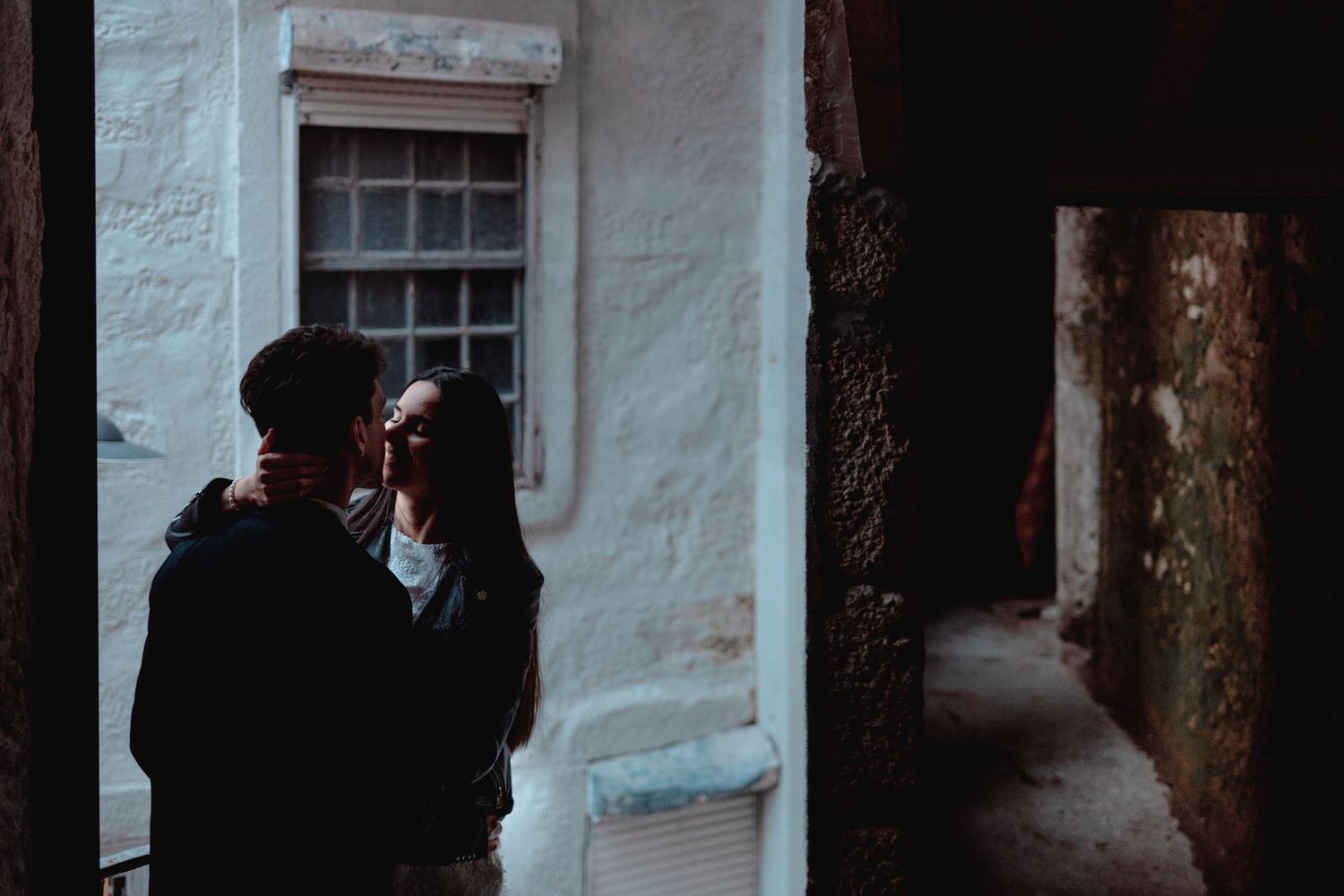 Romantic couple sharing an intimate moment in a moody alleyway beside a white stone building with vintage window.