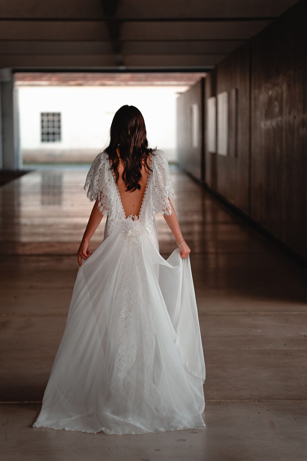V-back wedding dress with lace details photographed in dramatic hallway setting.