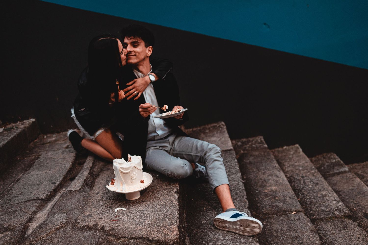 Couple sitting on outdoor steps sharing cake and drinks against a dark blue wall.