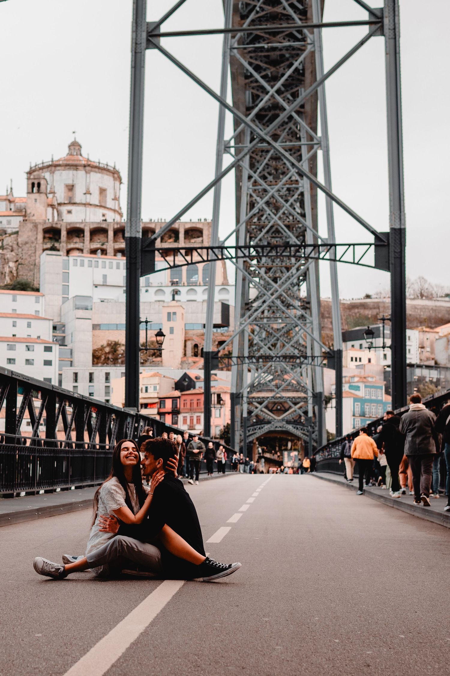 Couple sitting on Dom Luis I Bridge in Porto, Portugal, with historic buildings and iron lattice structure in background.