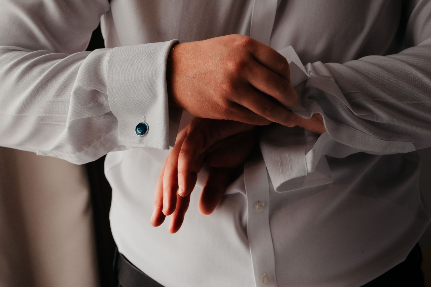 Man in white dress shirt fastening teal cufflinks, close-up of hands and French cuffs.