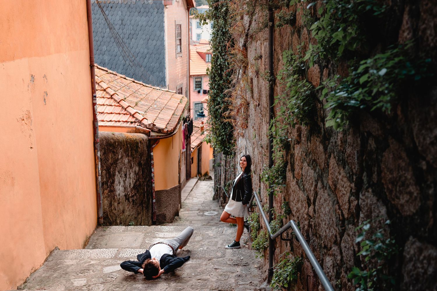 Photographer lying on cobblestone alley steps capturing woman in dress beside stone wall in historic European town.