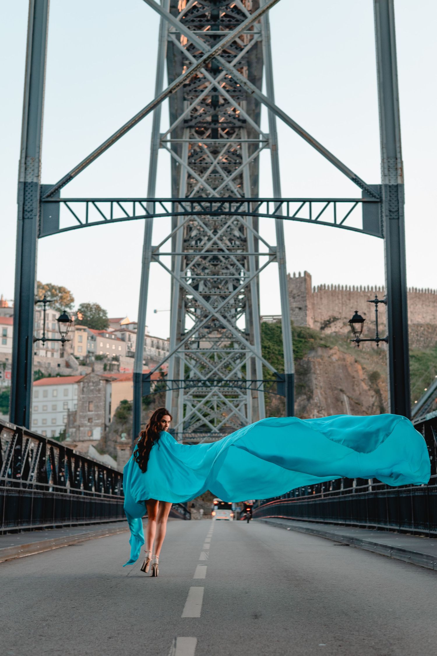 Woman in flowing turquoise dress poses dramatically on Porto's iconic bridge with castle view.