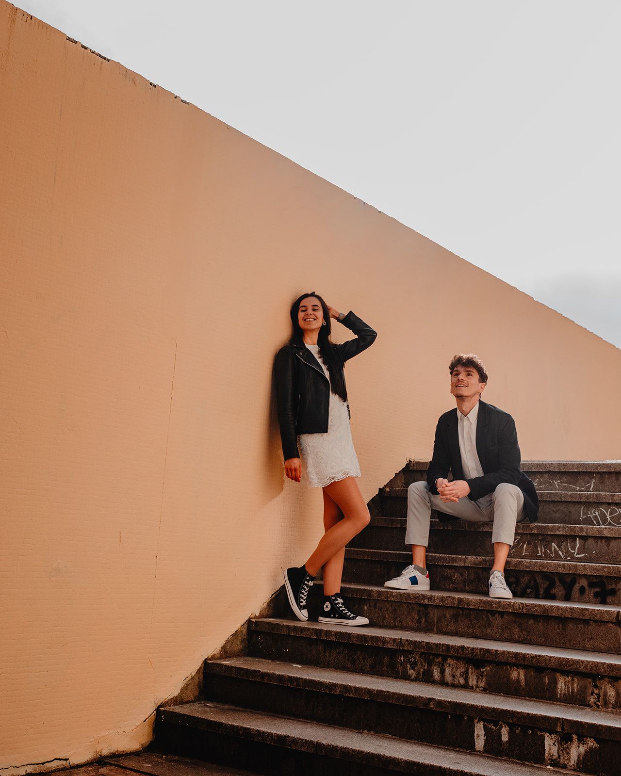 Couple sitting on outdoor concrete steps against peachy-orange wall at sunset.