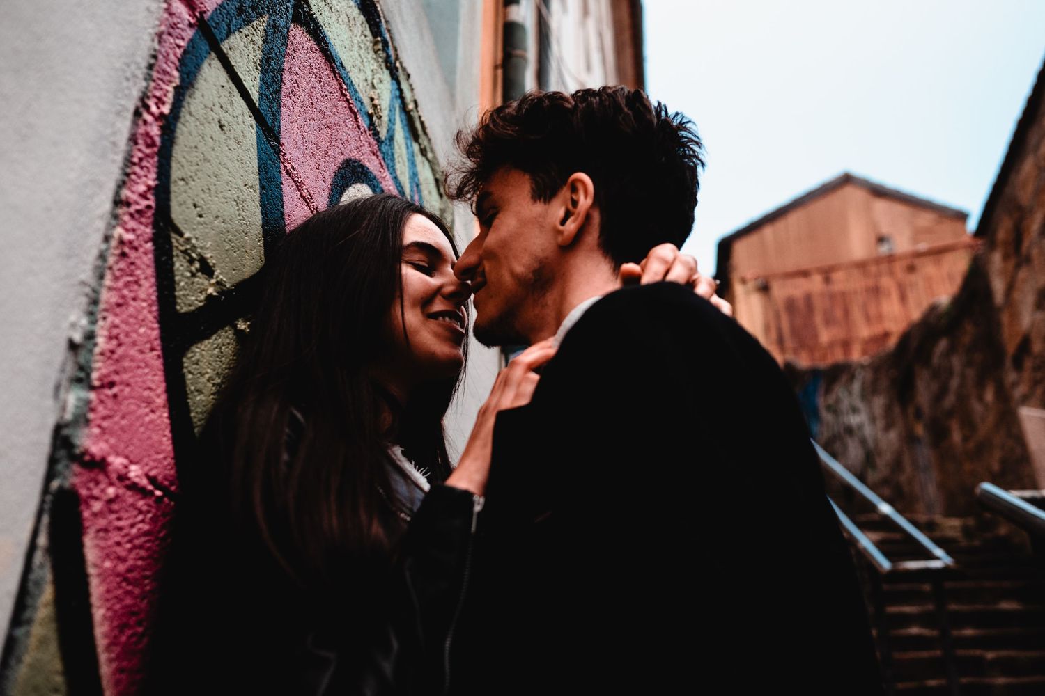 A young couple shares a romantic kiss against a colorful graffiti wall outdoors.
